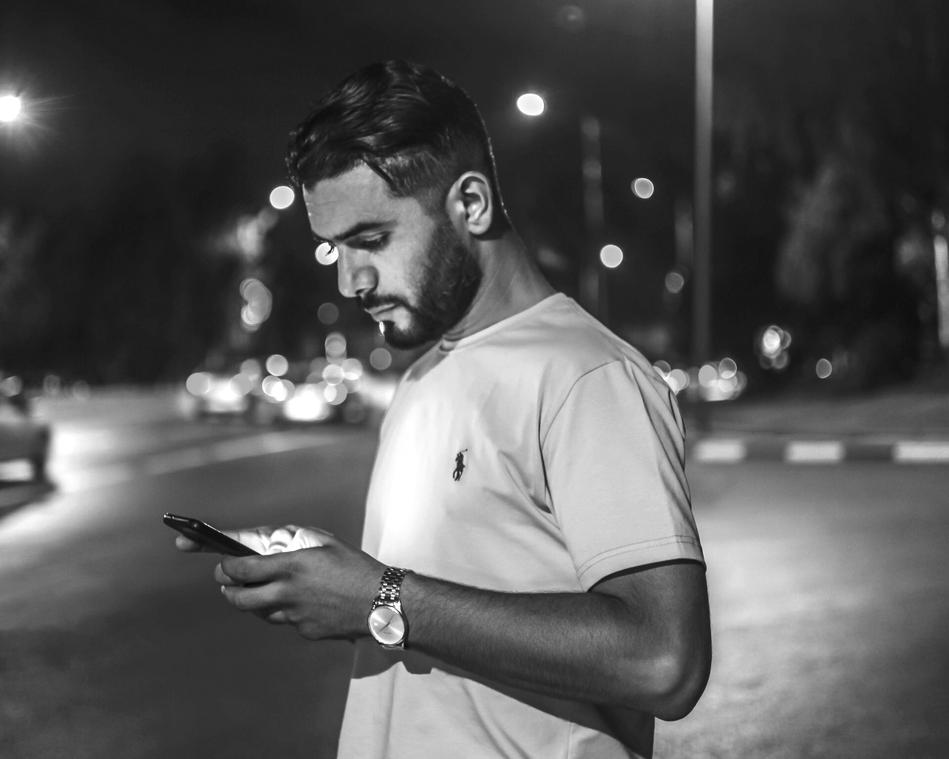 A stock image of a bearded man using a smartphone in public, standing on the side of the road as traffic passes in the background.