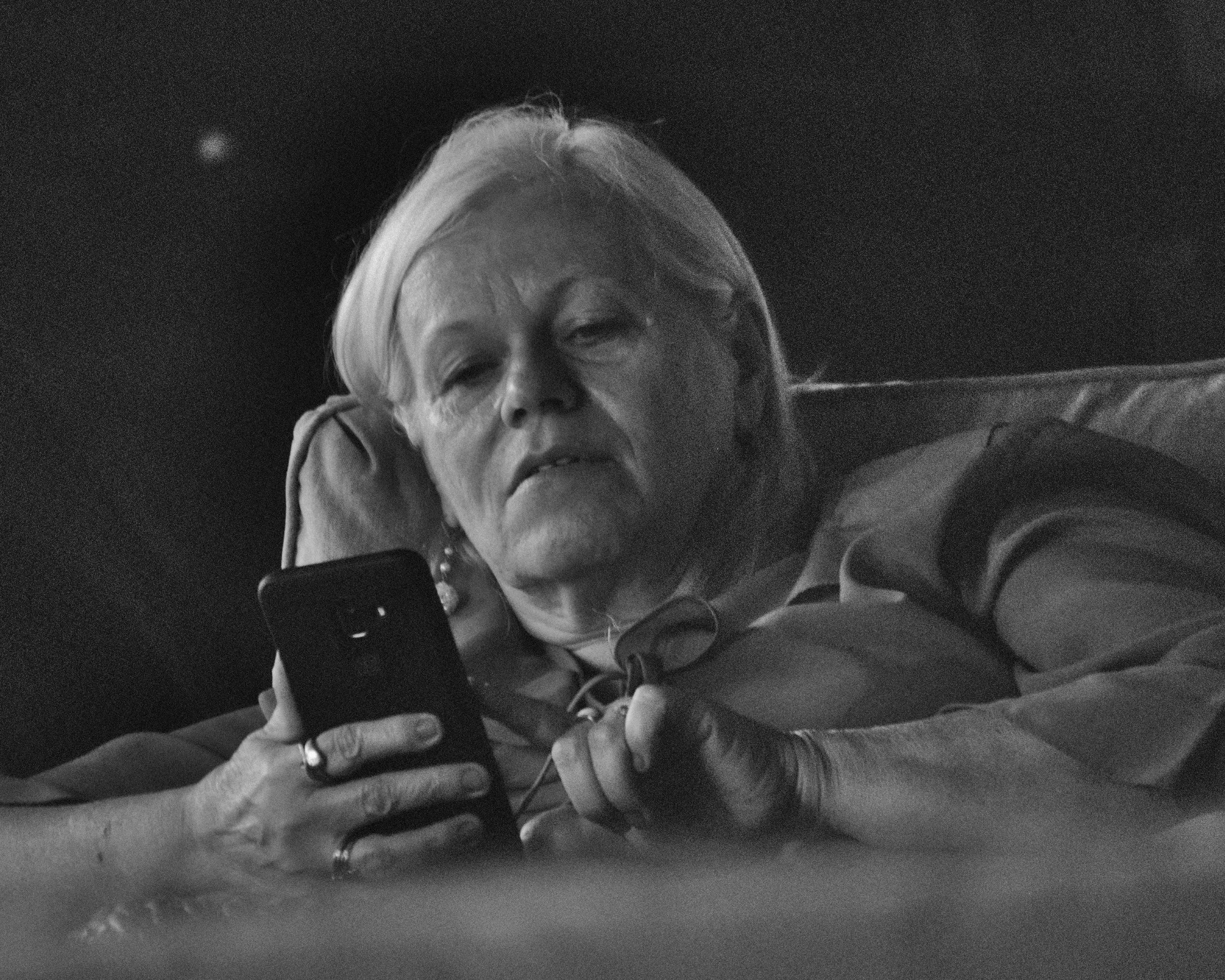 A stock image of a woman using a smartphone in low light from a reclining position, appearing to squint to get a better look at whatever she's reading on the device..