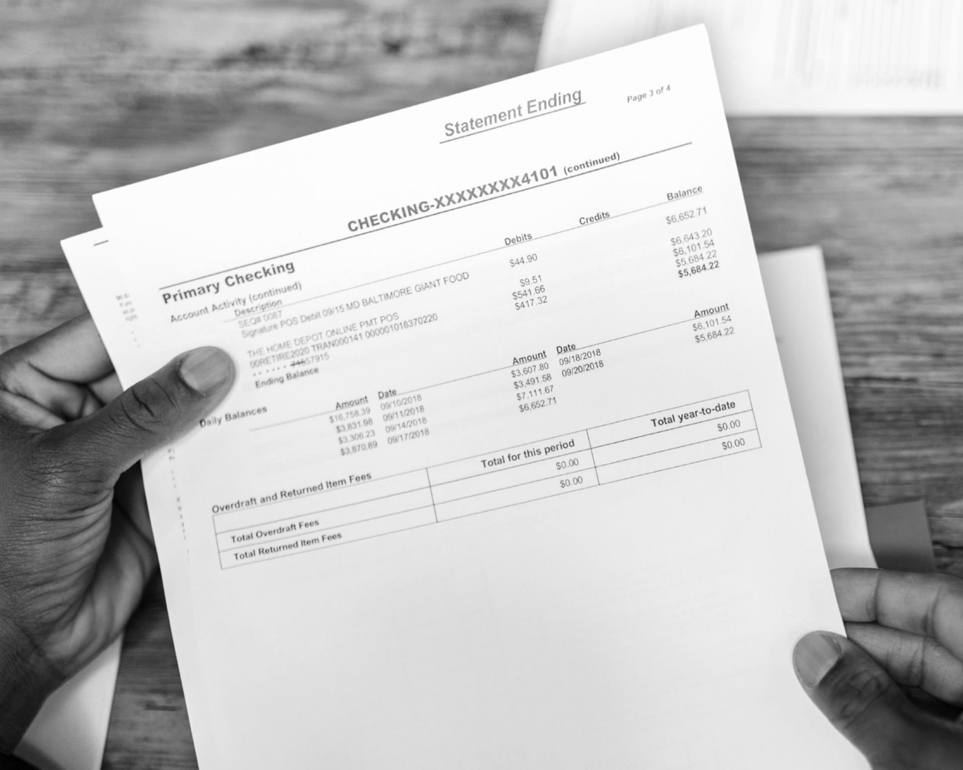 A stock image of a paper bank statement in a person's hands, positioned above a wooden desk with other papers placed on the desk.